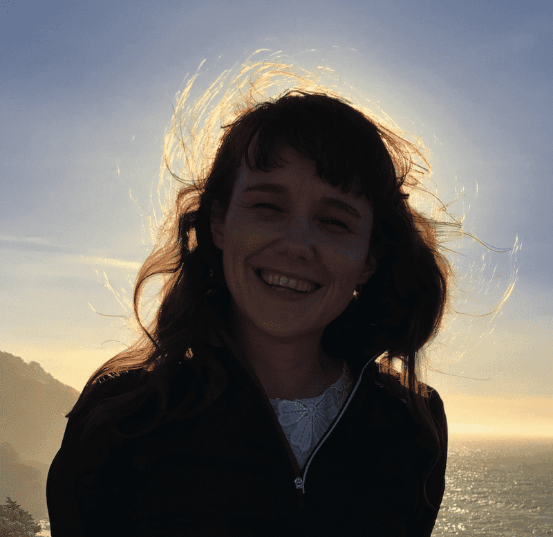 A photo of marie, backlit by the sun and ocean and grinning so wide their eyes are closed. The wind is making a mess of their hair. It's a bit hard to see their face, you just see that they are happy in the moment